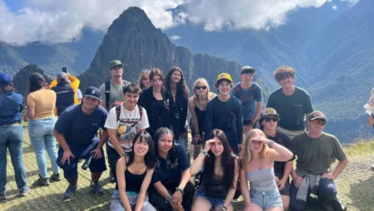 A group of people posing on a grassy area with mountains and a rocky peak in the background under a cloudy sky.
