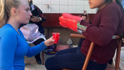 Two children engaged in craft activity; the child in the chair holds papier-mâché gloves, while the other kneels, holding a red brush. Colorful mural in the background.