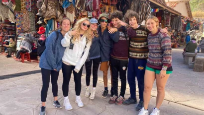 A group of seven young people in casual clothing stand together smiling in a vibrant outdoor market with textile goods on display.