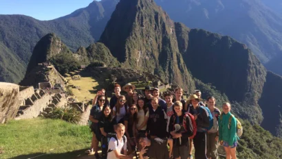 A group of people pose for a photo at the ancient Incan site of Machu Picchu, with the iconic mountain and ruins visible in the background under a clear blue sky.