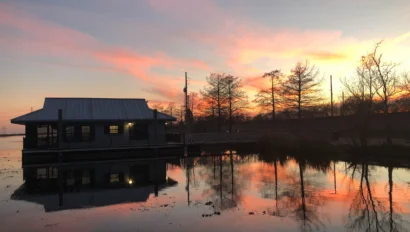 A small building sits by a calm lake during sunset, with trees and vibrant orange and pink clouds reflecting on the water's surface.