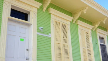 A green and yellow wooden house with decorative trim and closed shutters under a clear blue sky.