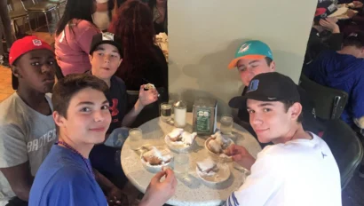 Five young people sit around a table eating beignets at a cafe. Each person wears a cap.