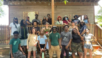 A group of people posing on wooden steps in front of a building, with trees in the background. Some are standing, and a few are seated, with various casual outfits and expressions.