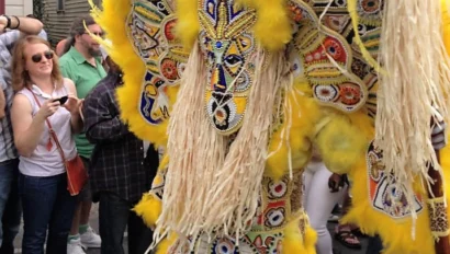 Person in a colorful, ornate costume with feathers and beads stands on a street, surrounded by onlookers.