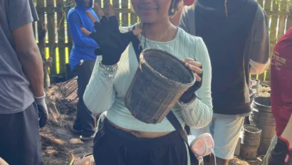 Person wearing a hat and gloves holds an empty pot while standing outdoors with a group of people.
