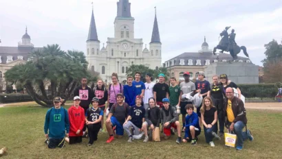 A group of people poses in front of St. Louis Cathedral and an equestrian statue, surrounded by greenery and a cloudy sky.