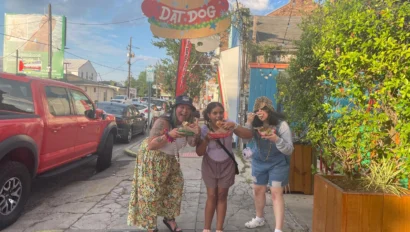 Three people enjoying hot dogs on a lively street outside a restaurant called "Dat Dog.