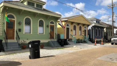 A row of colorful houses with arched windows and front steps line a street with utility poles and a parked truck under a clear blue sky. Trash bins and a traffic cone are on the sidewalk.