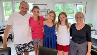 A group of five people, including three girls and two adults, standing together in a bright room with windows and a view of greenery outside.