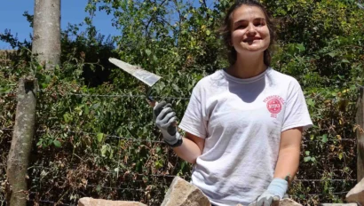 A person wearing gloves and a white T-shirt is smiling while holding a trowel, standing behind a partially constructed stone wall in a wooded area.