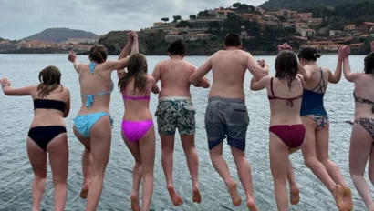 A group of people in swimwear are jumping into the water from a rocky ledge, with hills and buildings visible in the background.
