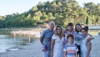 A group of six people smile on a rocky beach, with water and a forested hillside in the background.