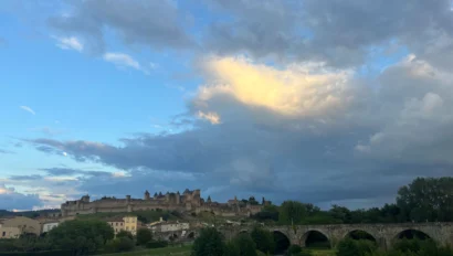 A scenic view of Carcassonne with a medieval fortress, houses, lush greenery, a river, and a stone bridge under a partly cloudy sky.