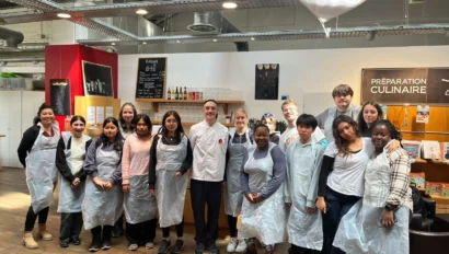A group of people wearing aprons stand together in a cooking class setting. Some kitchen equipment and culinary signs are visible in the background.