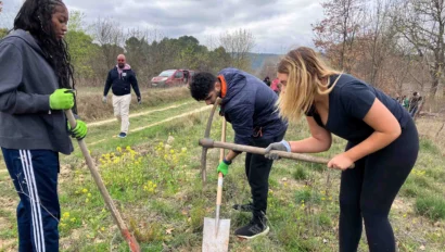 Three people use shovels and tools in a grassy, open field, while two others stand nearby. Overcast sky.
