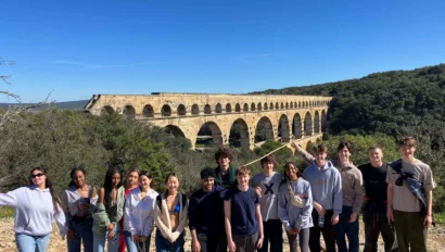 A group of people stands in front of the Pont du Gard aqueduct on a sunny day, with trees and blue sky in the background.