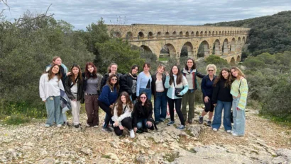 A group of people posing in front of an ancient stone aqueduct surrounded by greenery and a cloudy sky.
