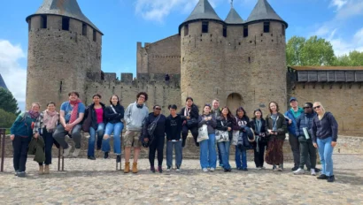 A group of people pose in front of a historic stone castle with round towers and conical roofs on a cobblestone plaza. The sky is partly cloudy.
