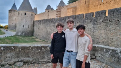 Three young men standing arm in arm on a stone walkway, with an ancient castle featuring round towers and high walls in the background.