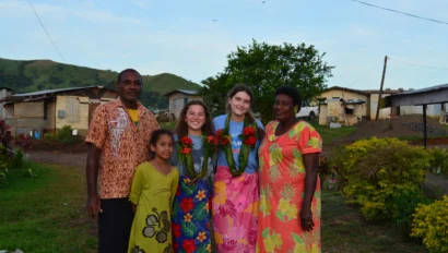 A group of six people, including children and adults, pose outdoors in front of rural houses and hills, wearing colorful clothing and smiling.