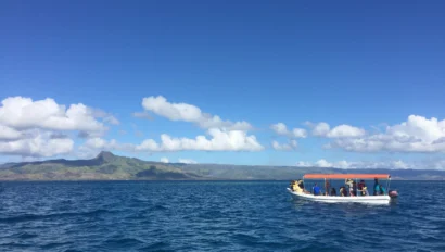A small boat with a canopy and several people on board floats on a vast body of water, with a mountainous landscape in the background under a clear blue sky.