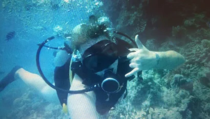 A scuba diver underwater, making a hand sign, with bubbles rising and coral visible in the background.