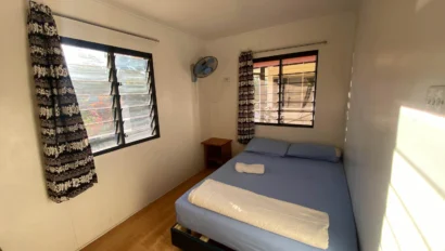 Small, sunlit bedroom with a single bed, blue bedding, a wall-mounted fan, two windows with patterned curtains, and a wooden nightstand.