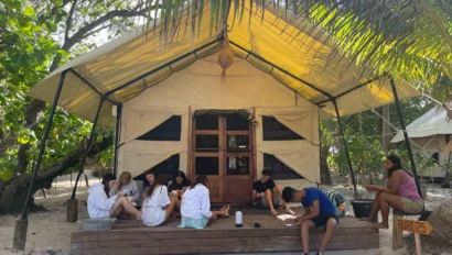 A group of people sitting on a wooden deck in front of a large tent surrounded by trees on a sandy ground. A sign reads "BURE 25.