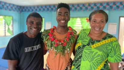 Three people stand indoors, smiling at the camera. The person in the center wears a floral garland, while the others wear brightly patterned clothing—capturing a joyful moment during a student travel program by Global Works.