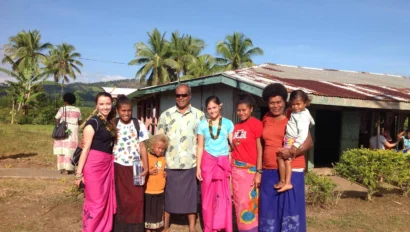 A group of people, including adults and children, stand smiling in front of a building in a tropical setting with palm trees. Some wear colorful traditional attire.