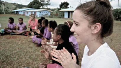 A group of children and an adult sit in a circle outdoors on grass clapping. The children wear purple uniforms and there are blue buildings in the background.