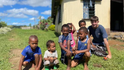 A group of children and a teenager sitting and squatting on grass in front of a wooden house, under a partly cloudy sky.