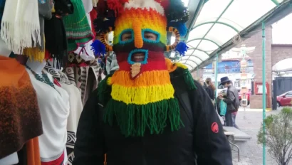 Person wearing a colorful, fringed mask in a marketplace, surrounded by hanging textiles.