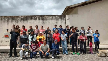 A group of people pose for a photo outdoors against a weathered wall. Some hold tools and a dog is present.