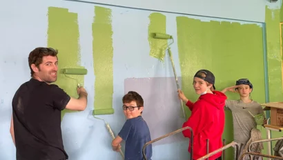 Four people painting a wall using rollers. The wall is partially painted green over a blue background. They are standing in casual clothing, engaged in the activity. Boxes are in the foreground.