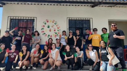 A group of people pose in front of a wall decorated with colorful handprints. Some hold up painted hands, and one person gives a thumbs-up.