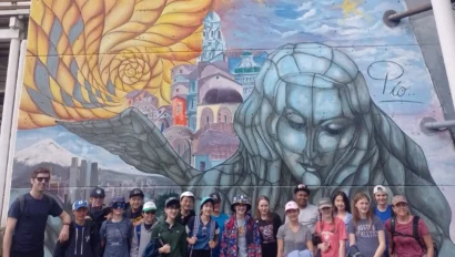 A group of people pose in front of a colorful mural featuring a large figure and buildings with a blend of abstract and realistic elements.