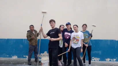 A group of six people holding paint rollers stand in front of a freshly painted wall, with a blue section at the bottom. They are posing for the photo and smiling.