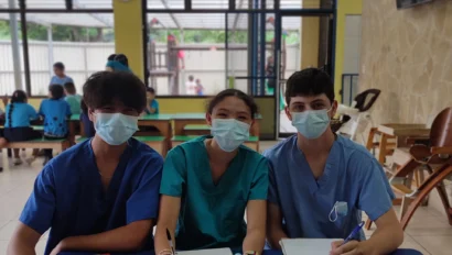 Three people in scrubs and masks sit at a table with notebooks and pens. They appear to be working or studying in a bright room with others in the background.