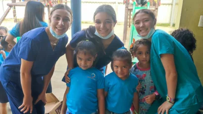 A group of adults and children posing together indoors near a play area. The adults are wearing scrubs, and the children are dressed casually.