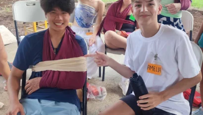 A group of young people sit outside. Two of them have arms in slings. One person holds a water bottle with a name tag reading "AMELIA." Trees are visible in the background.