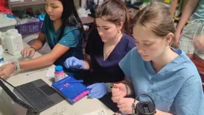 Three women in scrubs and gloves work at a table with medical supplies, paperwork, and a tablet, collaborating in a clinic setting as part of a pre med travel program.