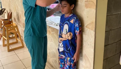 A healthcare worker, part of a pre med travel program, measures the height of a boy in Sonic pajamas against a wall using a stadiometer in a tiled room.