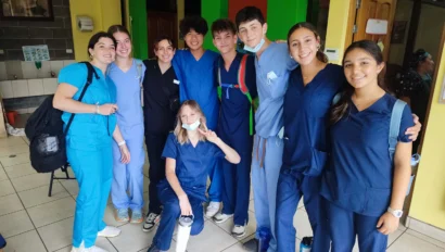 A group of nine young people in scrubs, part of a pre med travel program, pose together indoors, smiling at the camera. Some hold backpacks and water bottles, while one person flashes a peace sign.