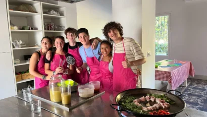 A group of six people wearing pink aprons stand together in a kitchen, smiling at the camera. A large pan with food is on the counter in front of them.