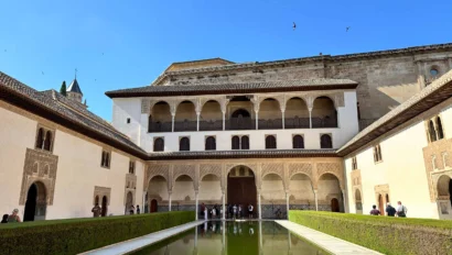 The image shows a historical courtyard with a reflective pool surrounded by a brown and white building. Arched balconies line the second floor, and birds are flying in the blue sky above.