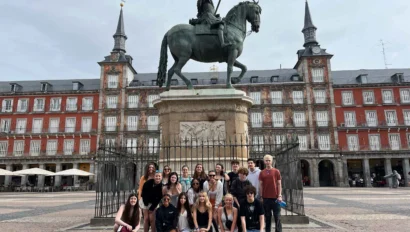A group of young people in the Spain summer travel program pose in front of an equestrian statue in the center of a large plaza with historic buildings in the background.