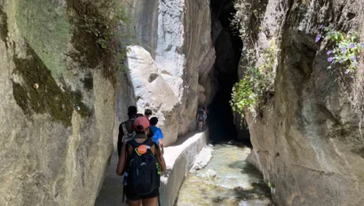People with backpacks hike along a narrow path beside a stream through a rocky canyon, enjoying sunlight and greenery above—an unforgettable experience on our Spain summer travel program.