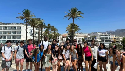 A group of people stands on a sunny plaza with palm trees and buildings in the background.